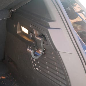 May include: A close-up of a black car interior, showcasing a metal hook attached to a dark panel. The hook is curved and appears to be designed for hanging items. A small light is visible in the background, illuminating the area.