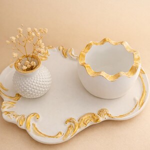 Könnte beinhalten: Ein weißes Keramik-Servier-Set mit goldenen Verzierungen. Das Set umfasst ein dekoratives Tablett, eine kleine Vase mit getrockneten Blumen und eine Schüssel. Das Tablett und die Schüssel haben einen goldfarbenen, gewellten Rand. Die Vase hat eine strukturierte Oberfläche.