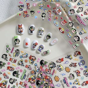 Op de afbeelding: Nail art stickers en geverfde nagels met de tekenfilmfiguren van de Powerpuff Girls. De stickers zitten op transparante vellen en de nagels zijn wit geschilderd met verschillende karakterontwerpen. De afbeelding bevat de tekst "Powerpuff Girls" en stickercodes.