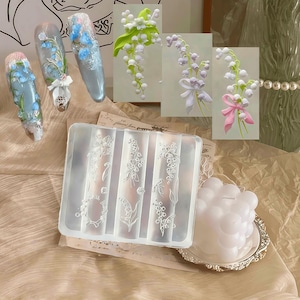 Può includere: Uno stampo in silicone trasparente con disegni floreali, insieme a tre nail art con accenti floreali blu e un fiocco bianco. Sono mostrate anche tre composizioni floreali di mughetto con foglie verdi e fiori bianchi, alcune con nastri.