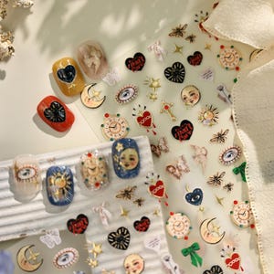 May include: A collection of nail art stickers and decorated nails. The stickers feature celestial and gothic designs, including moons, hearts, faces, and clocks. The nails are painted with intricate designs and embellishments, showcasing a variety of colors and textures.