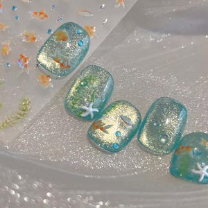 May include: A set of five artificial nails with a shimmering, light blue base. Each nail features a unique underwater design, including goldfish, starfish, bubbles, and aquatic plants. The nails are decorated with glitter and small, sparkling embellishments.