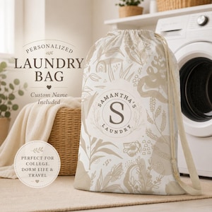 May include: A drawstring laundry bag featuring a beige and white botanical pattern. The center displays a circular emblem with the letter S and the text SAMANTHA'S LAUNDRY. The bag includes a drawstring closure and two shoulder straps for carrying.