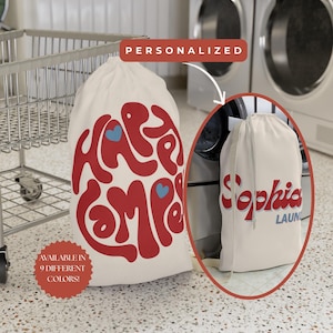 May include: Cream-colored laundry bag with a red, retro-style graphic that reads "Happy Camper" and two blue hearts. The bag has a drawstring closure. Text on the bag says "Available in 9 different colors!" and "Personalized".