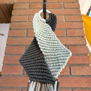 May include: A handmade macrame plant hanger in a diamond shape, featuring alternating sections of gray and light green knotted cord. The hanger is suspended from a black metal hook and white ring, with multiple cords extending downward.