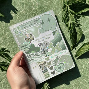 May include: A green and white Kindle cover decorated with stickers. The stickers feature a ghost, hearts, a butterfly, and text that says "hot girls read" and "a book a day keeps reality away."