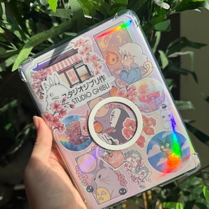 May include: A clear phone case with a pink background featuring Studio Ghibli anime stickers. The stickers include characters and imagery from various Ghibli films, with Japanese text. A white circular phone grip is attached.