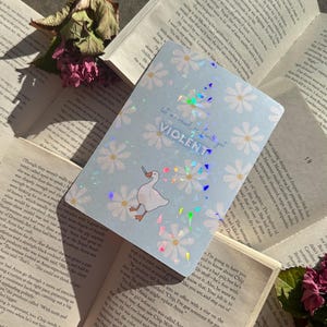 May include: A pale blue card featuring white daisy motifs and the text "let me read, or I will get VIOLENT". A cartoon goose is depicted. The card is set amongst open books and dried flowers.