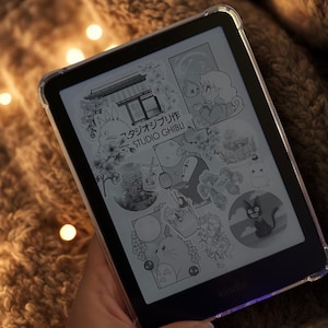 May include: A Kindle e-reader displaying black and white illustrations from Studio Ghibli films. The screen shows images of characters and scenes from various animated movies. The e-reader is in a clear protective case.