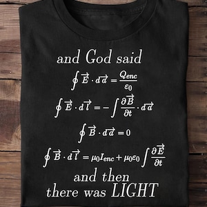 May include: A black t-shirt featuring white text and mathematical equations. The text reads: and God said, followed by Maxwell's equations, and then there was LIGHT. The design is centered on the front of the shirt with a crew neckline.