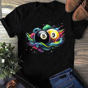 Puede incluir: Camiseta negra con un gráfico colorido de una bola 8 y 9 negras y amarillas. Las bolas están rodeadas de colores vibrantes y arremolinados. La camiseta se muestra con vaqueros azules y zapatos marrones.