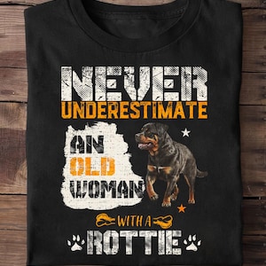 Könnte beinhalten: Schwarzes T-Shirt mit dem Text "NIEMALS EINE ALTE FRAU MIT EINEM ROTTIE UNTERSCHÄTZEN". Das Shirt zeigt eine Grafik eines Rottweiler-Hundes und Pfotenabdrücke. Der Text ist in Weiß und Orange gehalten.