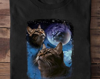 Pode incluir: Camiseta preta com um gráfico de três gatos olhando para uma lua cheia e um céu estrelado. Os gatos são tigrados marrons e cinzas, com pelos e bigodes detalhados. O fundo é um gradiente azul e preto.