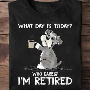 Op de afbeelding: Zwart T-shirt met een cartoon Schnauzer hond die een koffiemok vasthoudt. De tekst luidt "WHAT DAY IS TODAY? WHO CARES? I'M RETIRED" in het wit. De hond is grijs en wit.