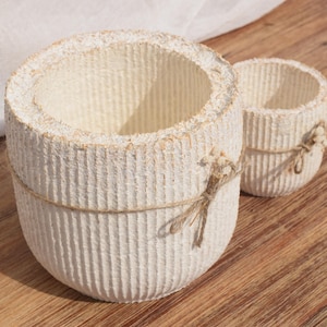 Peut inclure: Deux jardinières cylindriques texturées blanc cassé de différentes tailles. La plus grande a une bande de ficelle au milieu et un nœud en ficelle. La plus petite a une bande et un nœud en ficelle. Les deux sont sur une surface en bois.