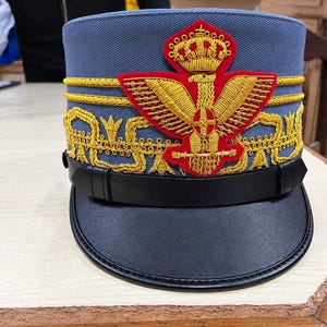 Puede incluir: Una gorra militar azul con un destacado emblema bordado en rojo y oro de un águila coronada con las alas extendidas. La gorra incluye adornos de trenza dorada, una correa de barbilla de cuero negro y una visera negra con bordes cosidos.