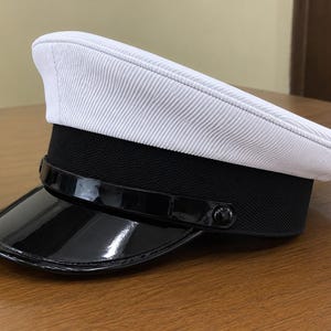 Op de afbeelding: Een witte pet met een klep, voorzien van een textuur op de witte bovenkant, een brede zwarte band en een glanzende zwarte lakleren klep. De pet heeft een dunne zwarte kinband die aan beide zijden is bevestigd met kleine zwarte knopen.