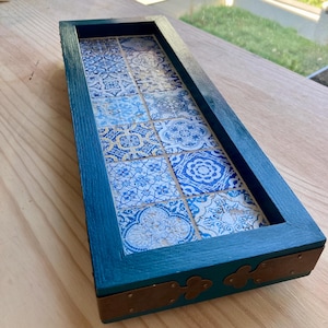 May include: A rectangular wooden tray with a teal painted frame. The tray's interior features a mosaic of blue and white patterned tiles. The corners are reinforced with bronze-coloured metal accents. The tray is displayed on a light-coloured wooden surface.