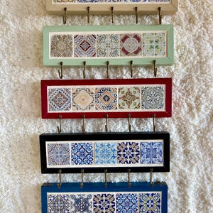 Pode incluir: Cinco organizadores de parede retangulares com molduras em bege, verde claro, vermelho, preto e azul. Cada organizador apresenta cinco azulejos decorativos com padrões geométricos complexos em azul, vermelho, verde e amarelo e cinco ganchos.
