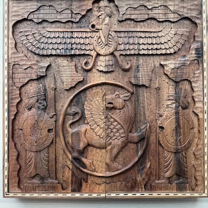 May include: A square wooden board featuring intricate relief carvings of a winged figure, a central winged lion within a circular frame, and two flanking figures holding spears and shields. The dark brown wood has a natural grain and a decorative border.