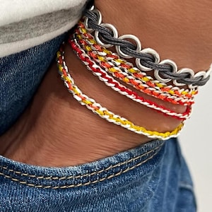 May include: A collection of colorful bracelets. The bracelets include a silver chain link bracelet and several braided bracelets in red, yellow, orange, and white. The bracelets are stacked on top of each other.