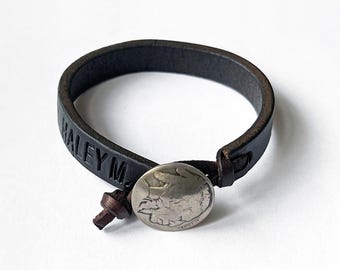 May include: A black leather bracelet with the name "Haley M." embossed on it. The bracelet has a silver coin charm with a design on it.