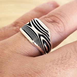 Pode incluir: Um anel de prata com uma face retangular com um padrão de listras pretas e brancas. O anel é usado em um dedo, mostrando seu metal polido e design exclusivo. A face do anel tem uma forma curva.