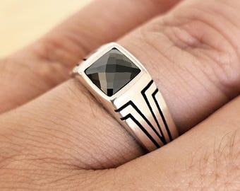 May include: A silver ring with a square, black gemstone. The ring has a geometric design on the band. The gemstone is set in a square bezel. The ring is shown on a person's finger.