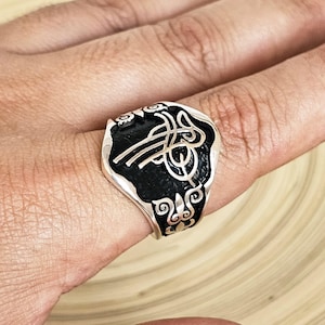 May include: A silver ring with a black enamel inlay featuring a stylized design. The ring has a rectangular face with rounded edges and ornate detailing along the sides. The design includes curved lines and geometric shapes.