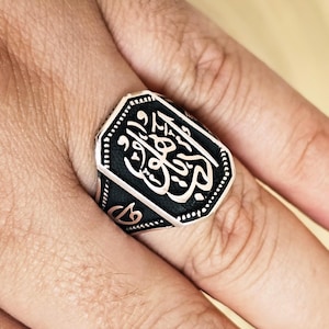 May include: A silver ring with a black rectangular face featuring white Arabic calligraphy. The ring has an octagonal shape and is worn on a finger. The ring's design is detailed and intricate.