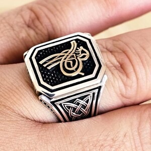 Puede incluir: Un anillo plateado con un diseño negro y dorado. El anillo presenta una cara cuadrada con un símbolo dorado adornado sobre un fondo negro. La banda tiene un diseño de nudo celta. El anillo está en un dedo.