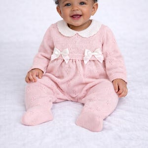 May include: A baby in a pink knit romper with a white collar and two white bows. The romper has long sleeves and attached feet. The baby is smiling and has dark curly hair.