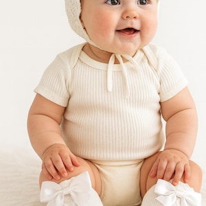 May include: A smiling baby wearing a cream-colored bonnet, short-sleeved onesie, and socks with white bows. The baby is sitting and looking towards the right side of the frame. The outfit is made of soft, ribbed fabric.