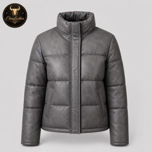 Könnte beinhalten: Graue Steppjacke mit hohem Kragen und Frontreißverschluss. Die Jacke hat ein gestepptes Design und zwei Seitentaschen. Das Ebon Leather-Logo ist oben links sichtbar.