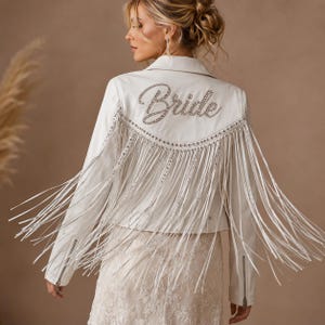 Könnte beinhalten: Weiße Lederjacke mit Fransen und dem Wort "Bride" in glitzernder Schrift auf dem Rücken. Die Jacke hat einen klassischen Biker-Stil mit Kragen, Reißverschluss und langen Ärmeln. Die Fransen sind silberfarben und hängen von den Schultern.