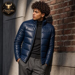 K&ouml;nnte beinhalten: Marineblaue Leder-Steppjacke mit Stehkragen und Rei&szlig;verschluss. Die Jacke hat ein gestepptes Design und wird &uuml;ber einem schwarzen Rollkragenpullover getragen. Das Ebon Leather-Logo befindet sich in der oberen linken Ecke.
