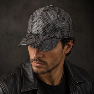May include: A dark gray and black baseball cap with a snakeskin pattern. The cap has a curved brim and a small button on top. The cap is made of a textured material and is worn by a person.