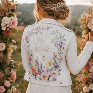 May include: White leather jacket with a floral design and the name "Mrs. Schmidt" in gold script. The jacket features a colourful array of flowers in pink, purple, yellow, and blue. A unique piece for a wedding or special occasion.