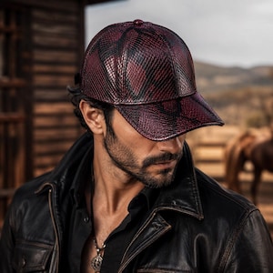 Puede incluir: Gorra de béisbol con estampado burdeos y negro. La gorra tiene una visera curva y una apariencia texturizada similar a la piel de serpiente. La gorra la lleva una persona que lleva una chaqueta de cuero negra.