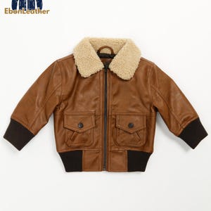 May include: A brown leather jacket with a beige shearling collar. The jacket has a zipper closure, two front pockets, and black ribbed cuffs and hem. The "EbonLeather" logo is in the upper left corner.