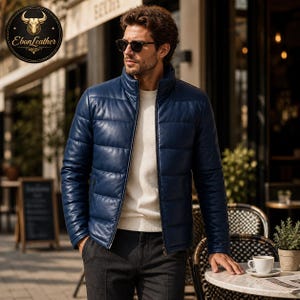 K&ouml;nnte beinhalten: Eine marineblaue Leder-Pufferjacke mit hohem Kragen und durchgehendem Rei&szlig;verschluss. Die Jacke wird &uuml;ber einem wei&szlig;en Pullover getragen. Das Bild zeigt auch eine Person mit Sonnenbrille und dunkler Hose. Die Jacke ist von EbonLeather.