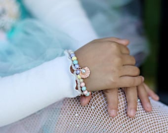 May include: A pastel beaded bracelet with a seashell charm. The bracelet features a mix of light blue, yellow, pink, and white beads. The charm is a pink seashell with a pearl in the center. The bracelet is on a child's wrist.
