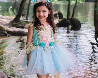 May include: A light blue and gold sleeveless dress featuring a sequined bodice, a layered tulle skirt, and floral accents at the waist. The outfit includes a matching headband adorned with teal flowers and a seashell-shaped embellishment.
