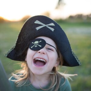 May include: A child wearing a black pirate hat with gold trim and a black eye patch with a skull and crossbones. The hat has a white crossbones design. The child is smiling with their mouth wide open.