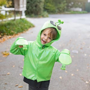 May include: A child wearing a bright green alien costume with a hood, sunglasses, and attached gloves. The costume has a shiny, metallic finish and a button closure. The child is smiling and appears to be outdoors.
