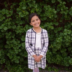 May include: A young person wearing a white and black plaid jacket and skirt set with a white top and pearl necklace. The outfit has black trim. The background is a wall of green leaves.