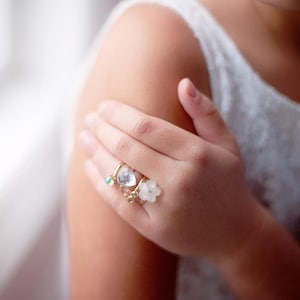 May include: A hand wearing four gold rings. The rings feature a small blue gem, a clear heart-shaped stone, a small gold bow, and a white flower. The rings are on a person's hand, with a white top in the background.