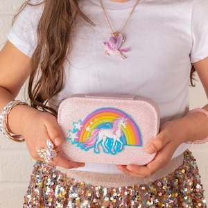 May include: A pink glittery jewelry box with a unicorn and rainbow design, held by a person. The person is also wearing a unicorn pendant necklace, a butterfly ring, and bracelets. The skirt is covered in sequins.
