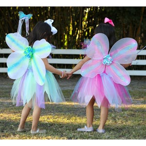May include: Two fairy costumes featuring layered tulle skirts in pastel shades of blue, green, and pink. Each outfit includes sheer, iridescent wings with a central floral accent and a matching headband. One costume is paired with a butterfly wand.