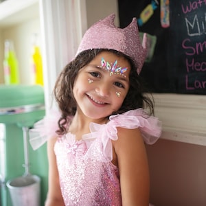 May include: A smiling child wearing a pink sequined dress, a matching crown, and face gems. The dress has tulle accents on the shoulders. The background includes a chalkboard and a vintage-style green appliance.